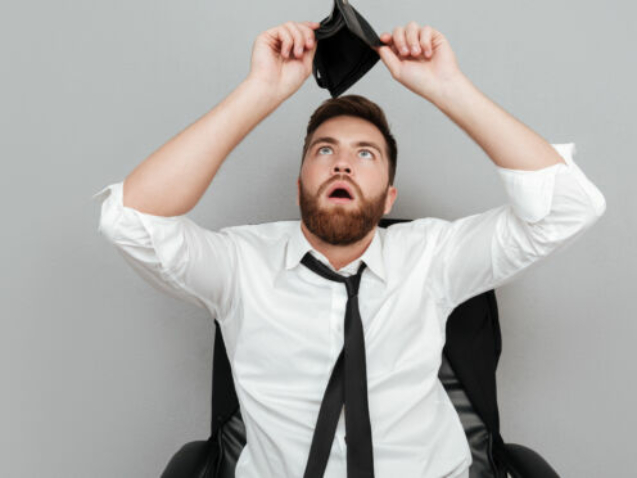 Frustrated surprised bearded man in white shirt showing empty wallet while sitting in chair isolated over gray background